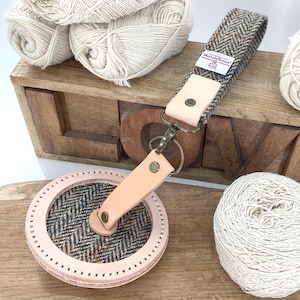 Puede incluir: Una colección de accesorios para manualidades, que incluye un ovillador de hilo rosa y tweed, un llavero de tweed y cuero, y varias madejas de lana blanca. Los artículos están dispuestos sobre una superficie de madera con la palabra "LOVE" tallada en una caja de madera.