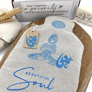 以下が含まれることがあります： 瞑想する人物の青いイラストと「Beautiful Soul」の文字が入ったベージュの湯たんぽ。ボトルには青いオムのシンボルが付いたタグが付いています。「Make happiness a priority」と書かれた箱が背景にあります。