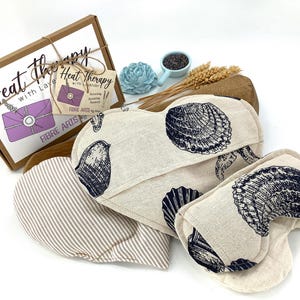 Peut inclure: Trois packs de thérapie thermique avec un motif de coquillages. Les packs sont faits de tissu naturel et remplis de graines de lin et de lavande. Les packs sont en forme de cœur et de rectangle long. Les packs sont emballés dans une boîte brune avec le texte "Heat Therapy with Lavender" sur le devant.