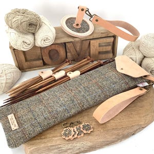 May include: A collection of knitting and crochet supplies, including yarn, needles, and a tweed case. The wooden block spells out "LOVE". The items are arranged on a wooden surface, with leather accents.