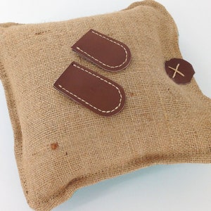 May include: A square burlap pillow with two brown leather patches and a brown leather button with an X stitched on it. The leather patches are stitched with white thread.
