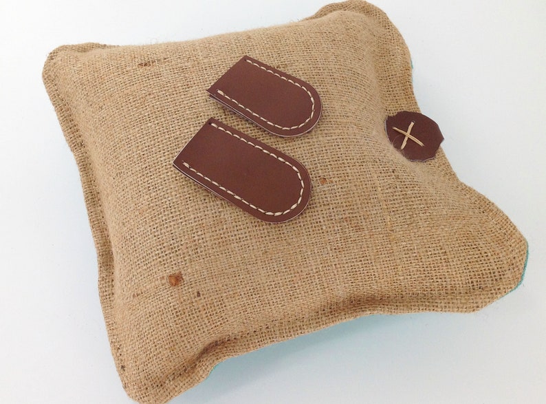 May include: A square burlap pillow with two brown leather patches and a brown leather button with an X stitched on it. The leather patches are stitched with white thread.