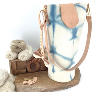 Puede incluir: Un bolso tote blanco y azul teñido anudado con una solapa y correa de cuero marrón claro. El bolso tiene herrajes dorados y está junto a una caja de madera con ovillos de lana y un alfiler decorativo.