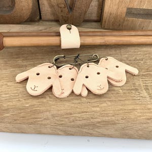 Può includere: Un set di quattro segnapunti a forma di pecora in pelle color cuoio chiaro, attaccati a una spilla da balia. Le pecore hanno semplici tratti del viso. Sono visibili anche ferri da maglia in legno.