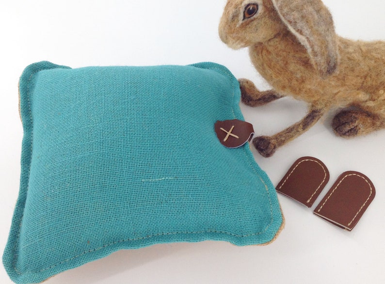 May include: A teal square pillow with a brown leather accent and a stitched 'X' detail. A felted brown rabbit and two brown leather thimble-shaped pieces are also in the image. The pillow has a beige border.