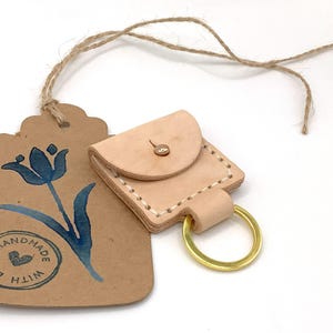 May include: A handmade leather keychain pouch with a gold-coloured ring. The pouch is light tan with a button closure. A brown tag with a blue tulip design and the words "LOVE HANDMADE WITH HIM" is attached.