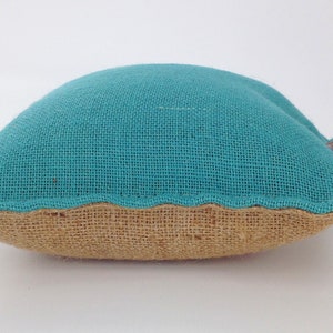 May include: A small, turquoise burlap pillow with a brown burlap base. The pillow has a curved shape and a small, brown leather loop attached to the top. The pillow is on a white background.