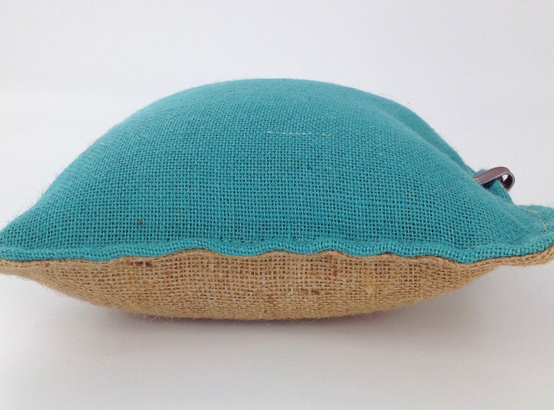 May include: A small, turquoise burlap pillow with a brown burlap base. The pillow has a curved shape and a small, brown leather loop attached to the top. The pillow is on a white background.