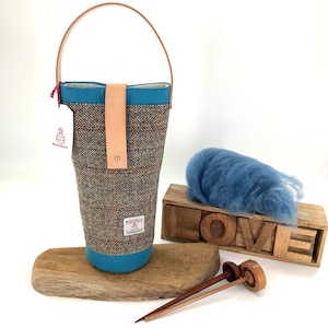 May include: A brown and blue tweed wine bottle carrier with a leather handle and strap. The carrier has a blue base and trim. A blue wool fiber, wooden block letters spelling "LOVE", and wooden knitting needles are also in the image.