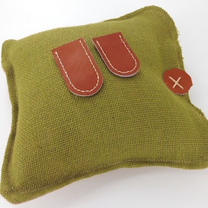 May include: A green burlap pillow with two brown leather straps and a brown leather button with an 'X' stitched on it.