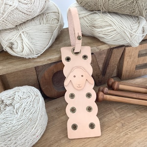 May include: A light pink leather sheep-shaped bookmark with metal eyelets and a loop. The bookmark is next to wooden knitting needles and balls of yarn. A wooden box with the word "LOVE" is in the background.