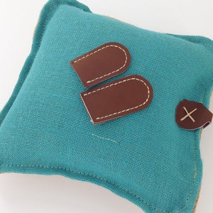 May include: A square teal linen pillow with brown leather accents. Two brown leather tags with white stitching are placed on the pillow, along with a leather button with an X-shaped stitch.