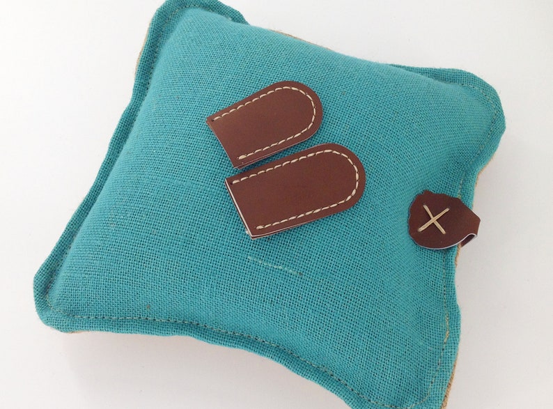 May include: A square teal linen pillow with brown leather accents. Two brown leather tags with white stitching are placed on the pillow, along with a leather button with an X-shaped stitch.