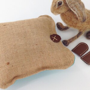 May include: A square burlap pillow with a brown leather accent and a felted hare figurine. Two small brown leather ear-shaped pieces are also present. The pillow has a turquoise lining.
