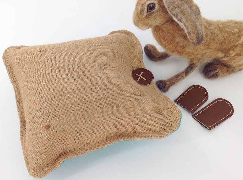 May include: A square burlap pillow with a brown leather accent and a felted hare figurine. Two small brown leather ear-shaped pieces are also present. The pillow has a turquoise lining.
