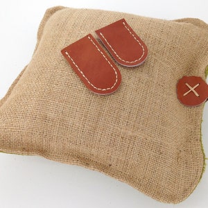 May include: A brown burlap pillow with two brown leather pockets and a brown leather strap with an X stitched on it.