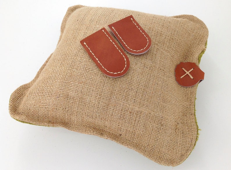 May include: A brown burlap pillow with two brown leather pockets and a brown leather strap with an X stitched on it.