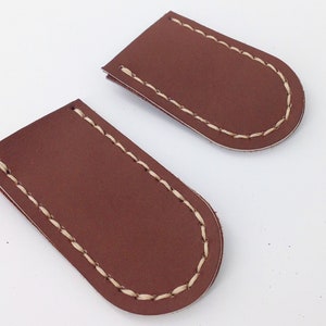 May include: Two brown leather pieces with rounded ends and cream-colored stitching. The leather has a smooth texture. The pieces are likely components of a larger item, possibly a wallet or a similar accessory.