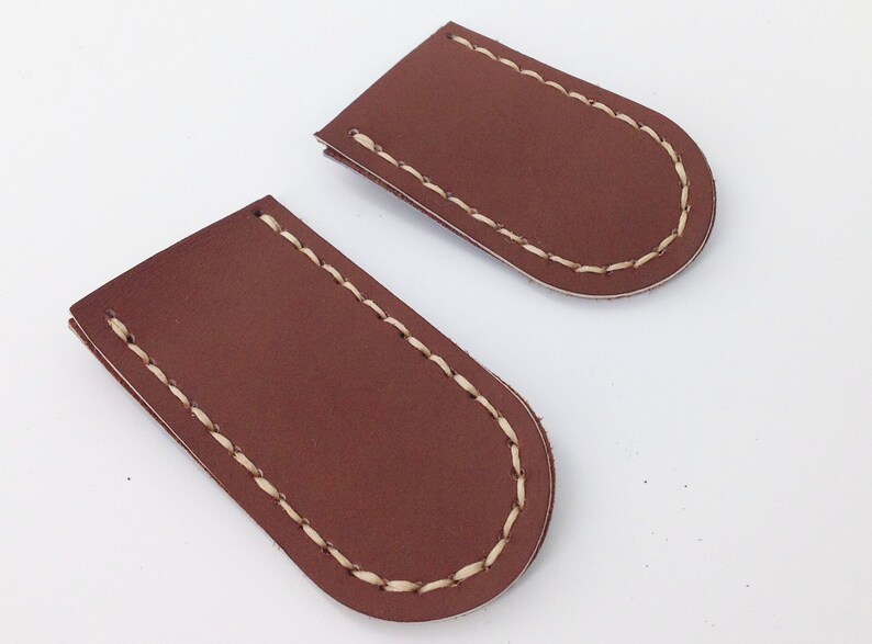 May include: Two brown leather pieces with rounded ends and cream-colored stitching. The leather has a smooth texture. The pieces are likely components of a larger item, possibly a wallet or a similar accessory.