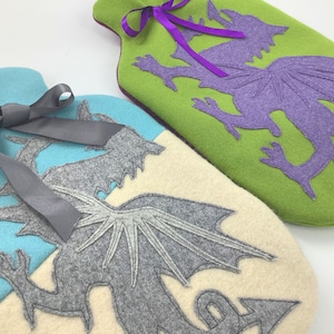 May include: Two felt hot water bottles with dragon designs. One bottle is green with a purple dragon, the other is blue with a gray dragon. Both bottles have a ribbon tied around the neck.