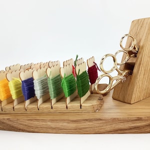 Puede incluir: Organizador de hilos de madera con una variedad de hilos de colores y un par de tijeras doradas. El organizador tiene ranuras para sujetar los hilos y un compartimento separado para las tijeras.