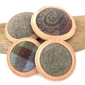 Può includere: Set di quattro sottobicchieri rotondi con centri in tessuto tweed in vari modelli e colori, tra cui verde, marrone e blu. Ogni sottobicchiere ha un bordo in pelle cucita. I sottobicchieri sono esposti su una superficie di legno.
