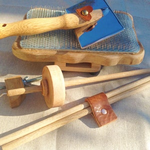 Puede incluir: Herramientas de madera para la preparación de fibra, incluyendo una carda con dientes metálicos y mango de madera. Una herramienta de metal azul con mango de madera y un acento de corazón de cuero. También se ve un huso de madera.