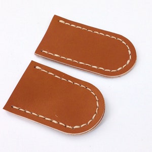 May include: Two brown leather patches with white stitching around the edges. The patches are shaped like rounded rectangles.
