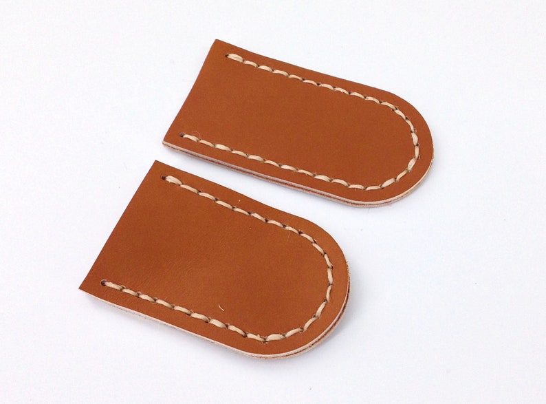 May include: Two brown leather patches with white stitching around the edges. The patches are shaped like rounded rectangles.