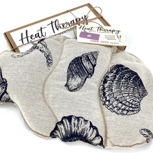 May include: A beige linen heat therapy pack with a seashell design. The pack is filled with lavender and flax seeds. The pack is packaged in a brown box with the text "Heat Therapy with Lavender" on it.