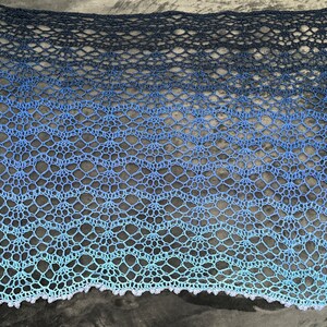 May include: A crocheted shawl with a gradient of blue colors, from dark blue to light blue. The shawl has a delicate lace pattern and a scalloped edge.