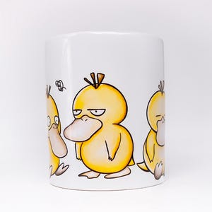 May include: White ceramic mug with a cartoon illustration of three yellow duck-like creatures with black outlines. The central character has a stern expression. The mug is cylindrical and has a glossy finish.
