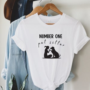 May include: White t-shirt with the text "NUMBER ONE pet sitter" in black script. A black silhouette graphic shows a person and a dog touching paws. The shirt is hanging on a wooden hanger.