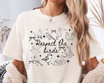 May include: Cream-colored t-shirt featuring a black and white illustration of various birds and the text "Respect the birds." The design includes stars and bows, creating a whimsical and nature-inspired aesthetic.