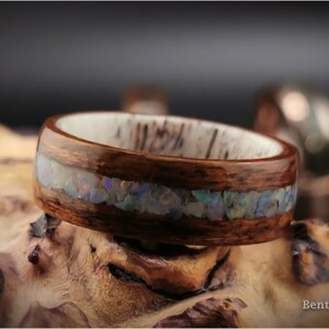 May include: A wooden ring with a band of opal inlay. The ring is made of dark brown wood and has a white inner band. The opal inlay is a thin line of iridescent stones.