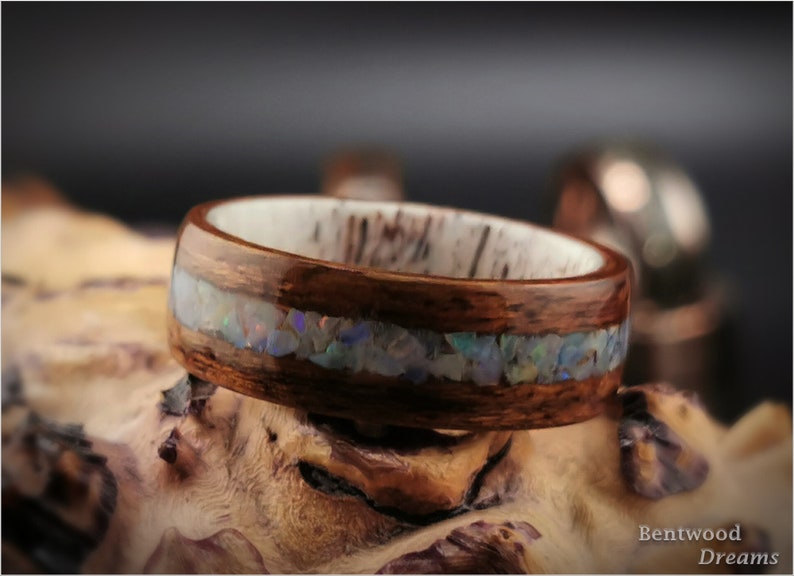 May include: A wooden ring with a band of opal inlay. The ring is made of dark brown wood and has a white inner band. The opal inlay is a thin line of iridescent stones.