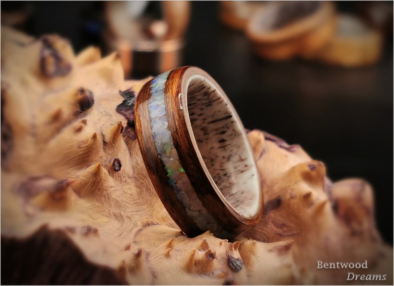 May include: A wooden ring with a band of opal inlay. The ring is set on a piece of wood with a natural grain pattern.