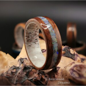 May include: A wooden ring with a band of white wood and a thin strip of opal inlay. The ring is on a piece of wood with the text "Bentwood Dreams".