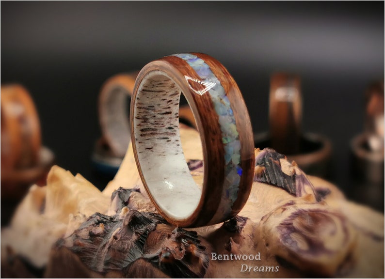May include: A wooden ring with a band of white wood and a thin strip of opal inlay. The ring is on a piece of wood with the text "Bentwood Dreams".