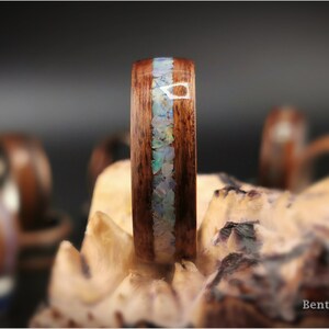 May include: A close-up of a wooden ring with a band of iridescent opal inlay. The ring is set on a piece of wood with the text "Bentwood Dreams" visible in the lower right corner.
