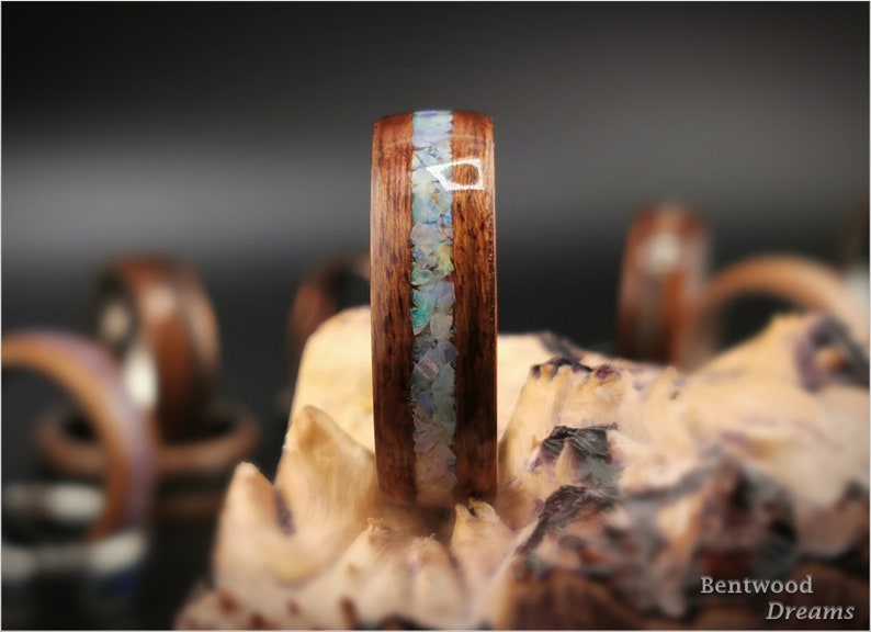 May include: A close-up of a wooden ring with a band of iridescent opal inlay. The ring is set on a piece of wood with the text "Bentwood Dreams" visible in the lower right corner.