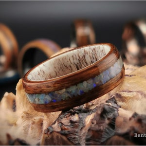 May include: A wooden ring with a band of brown wood and a thin inlay of iridescent blue and white opal. The ring is sitting on a piece of wood.