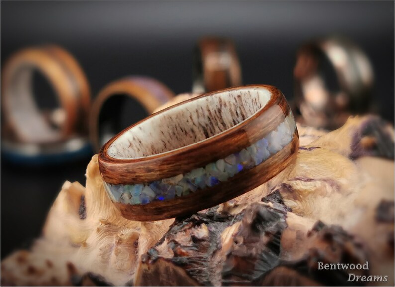 May include: A wooden ring with a band of brown wood and a thin inlay of iridescent blue and white opal. The ring is sitting on a piece of wood.