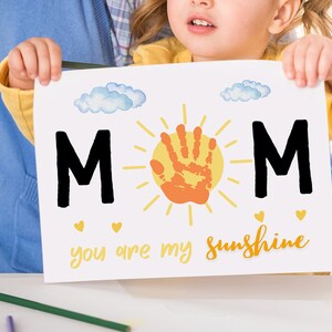 May include: A child holds up a white card with the word "MOM" in black letters, a handprint sun, and the text "you are my sunshine" in yellow.