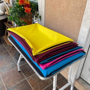 Pode incluir: Uma pilha de camas coloridas para animais de estimação em tons de amarelo, bordô, verde-azulado, rosa e azul. As camas são retangulares e parecem ser feitas de um material macio e acolchoado. Estão empilhadas numa mesa pequena.