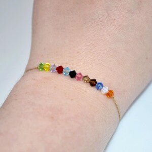 A close-up of a wrist wearing a Taylor Swift Eras Tour inspired bracelet. The bracelet features 12 crystal beads strung on a thin gold cord, each color symbolizing an album era. The beads are arranged in this order: green (Debut), yellow (Fearless), purple (Speak Now), red (Red), light blue (1989), black (Reputation), pink (Lover), tan (Folklore), brown (Evermore), dark blue (Midnights), white (The Tortured Poet's Department), and orange (The Life of a Showgirl).