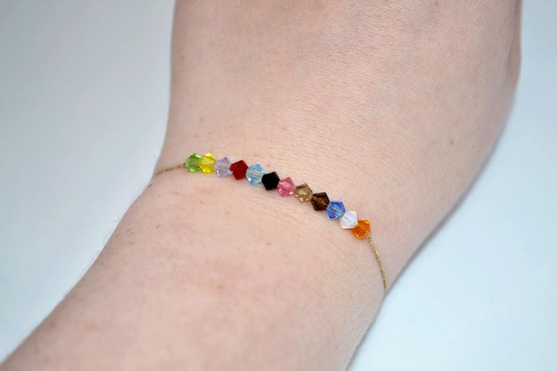 A close-up of a wrist wearing a Taylor Swift Eras Tour inspired bracelet. The bracelet features 12 crystal beads strung on a thin gold cord, each color symbolizing an album era. The beads are arranged in this order: green (Debut), yellow (Fearless), purple (Speak Now), red (Red), light blue (1989), black (Reputation), pink (Lover), tan (Folklore), brown (Evermore), dark blue (Midnights), white (The Tortured Poet's Department), and orange (The Life of a Showgirl).