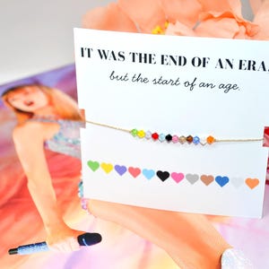 A Taylor Swift Eras inspired bracelet on a card that reads “IT WAS THE END OF AN ERA, but the start of an age.” The bracelet has 12 colorful crystal beads on a gold cord, each representing an era. Below, 12 hearts label the eras: green (Debut), yellow (Fearless), purple (Speak Now), red (Red), light blue (1989), black (Reputation), pink (Lover), tan (Folklore), brown (Evermore), dark blue (Midnights), white (The Tortured Poets Department), and orange (The Life of a Showgirl).