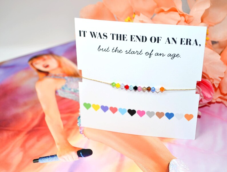 A Taylor Swift Eras inspired bracelet on a card that reads “IT WAS THE END OF AN ERA, but the start of an age.” The bracelet has 12 colorful crystal beads on a gold cord, each representing an era. Below, 12 hearts label the eras: green (Debut), yellow (Fearless), purple (Speak Now), red (Red), light blue (1989), black (Reputation), pink (Lover), tan (Folklore), brown (Evermore), dark blue (Midnights), white (The Tortured Poets Department), and orange (The Life of a Showgirl).