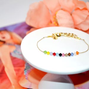 A Taylor Swift Eras inspired bracelet displayed on a white pedestal with peach flowers in the background and the Taylor Swift Eras Tour book. The bracelet features a thin gold cord and 12 colorful crystal beads, each representing an era: green (Debut), yellow (Fearless), purple (Speak Now), red (Red), light blue (1989), black (Reputation), pink (Lover), tan (Folklore), brown (Evermore), dark blue (Midnights), white (The Tortured Poets Department), and orange (The Life of a Showgirl).